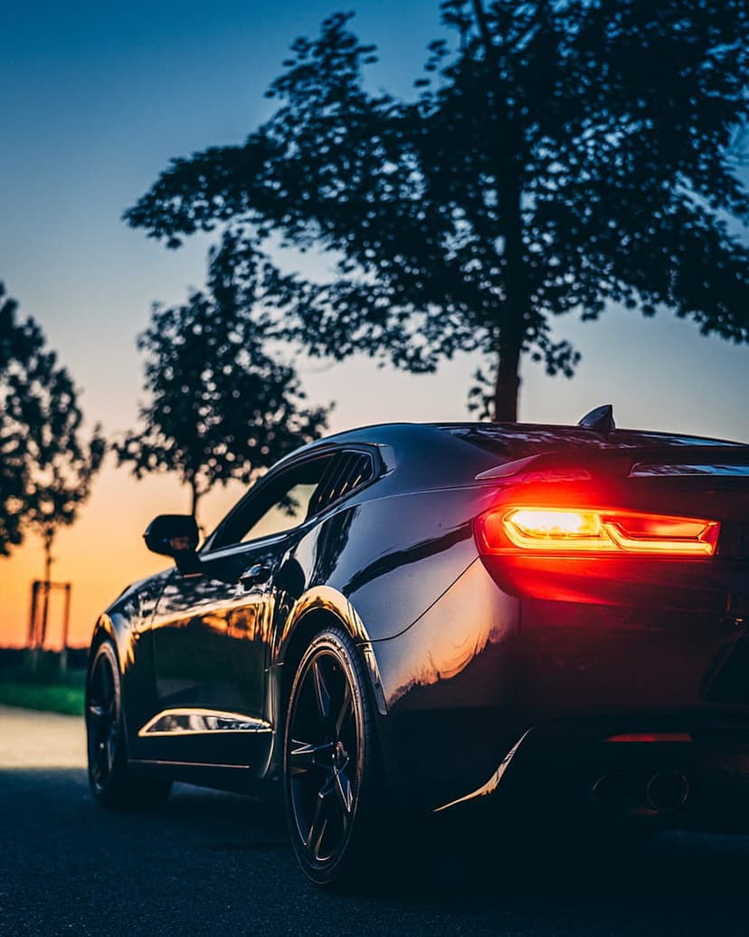 Black Chevrolet Camaro with glowing red taillights at sunset under a gradient orange and blue twilight sky.
