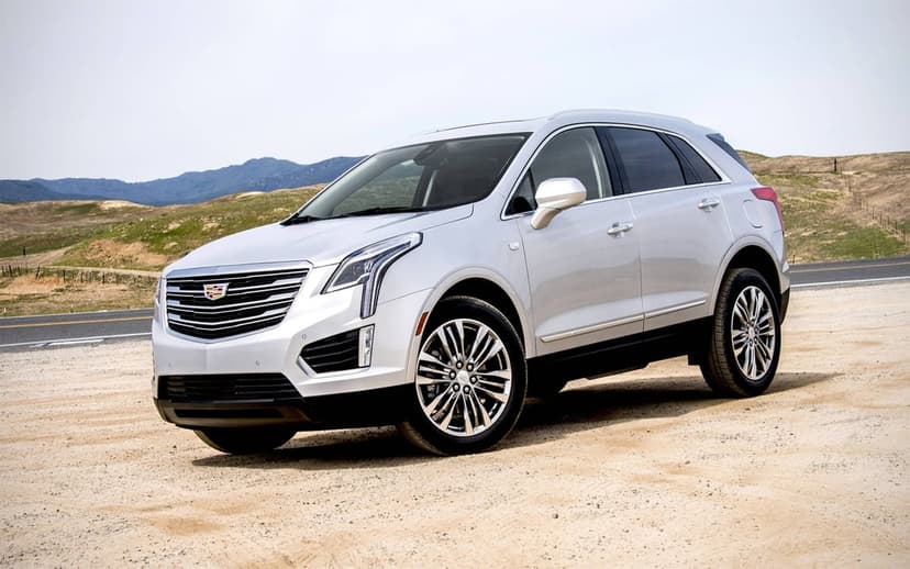 Silver Cadillac SUV parked on a sandy path with rolling green hills and blue mountains in the background.
