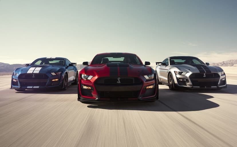 Three Ford Mustang Shelby GT500 cars in blue, red, and silver driving fast on a desert salt flat under a clear sky.