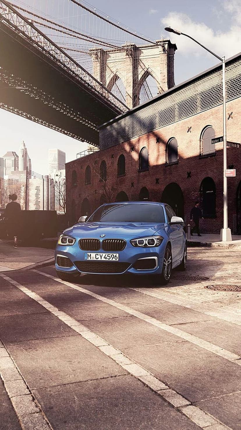Blue BMW sedan parked on a cobblestone street with the Brooklyn Bridge and brick buildings in the background.