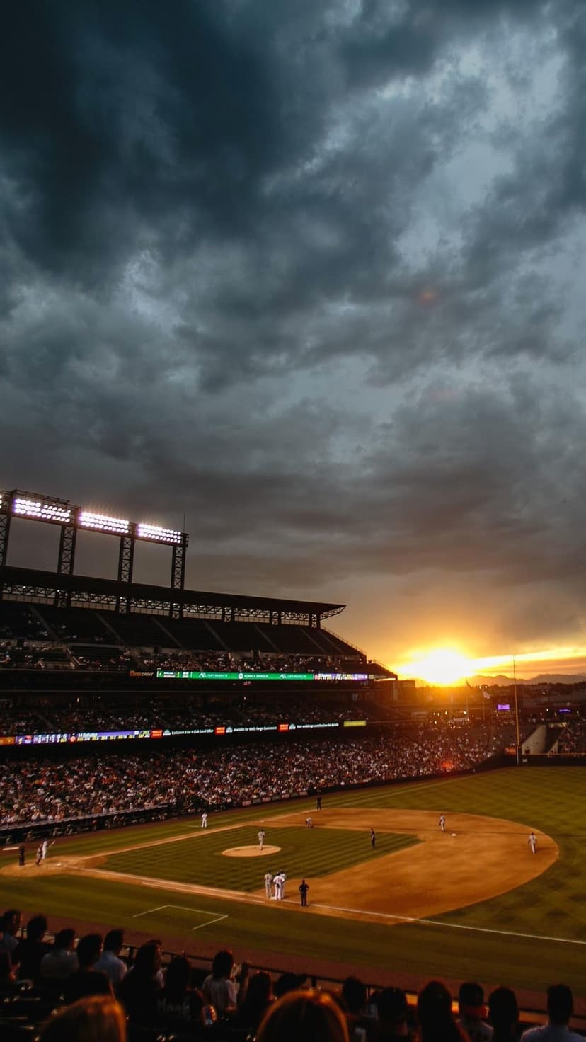 Baseball stadium under a fiery orange sunset with dark storm clouds and bright stadium lights over a green field.