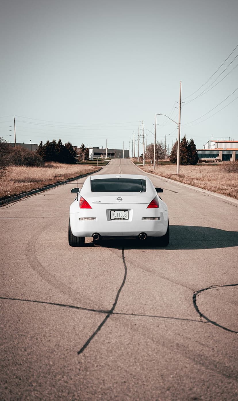 White Nissan 350Z parked on asphalt with black tire marks near dry grass and industrial utility poles.