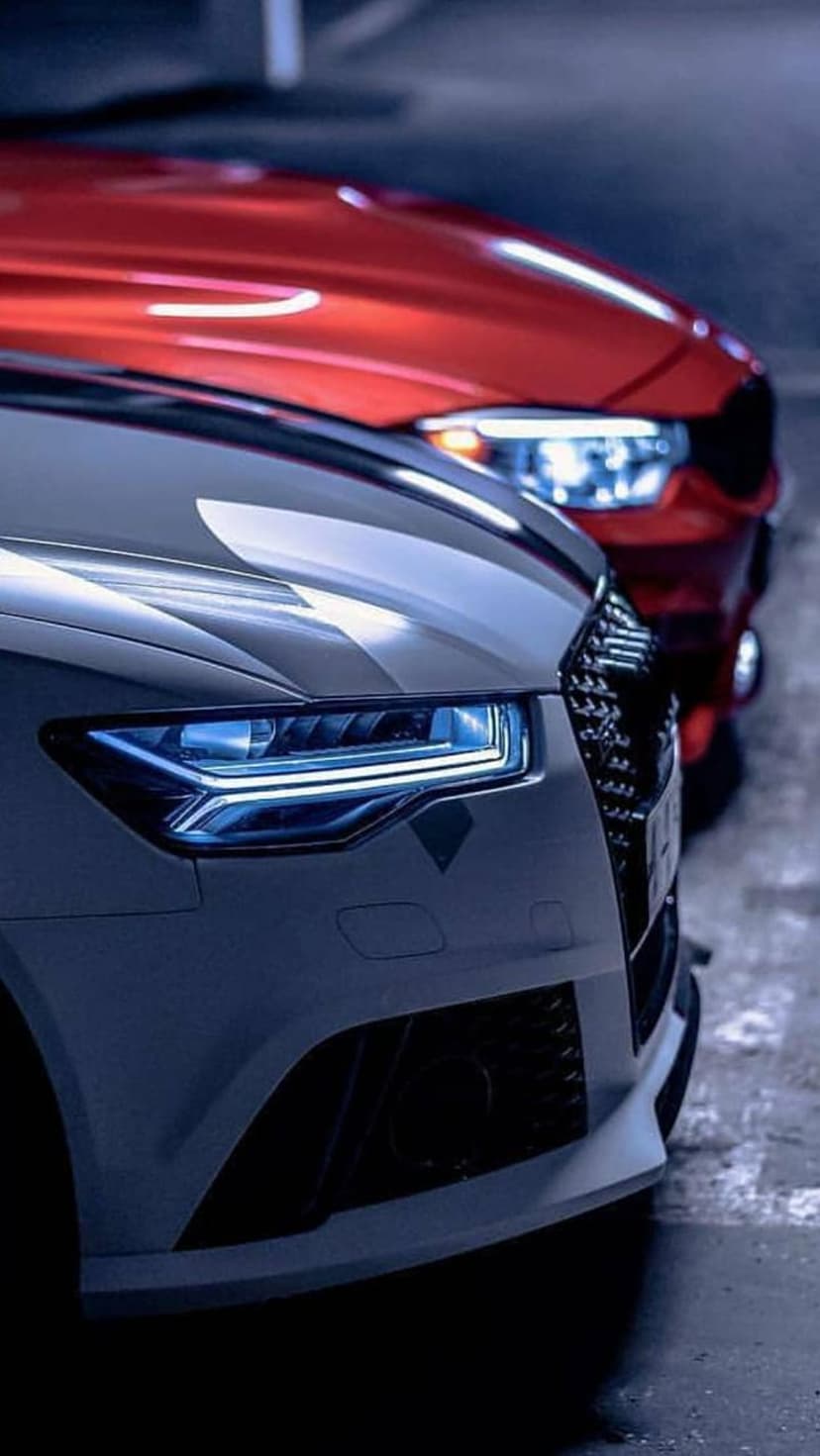 Grey Audi with illuminated headlights parked next to a red BMW in a dark concrete parking garage.
