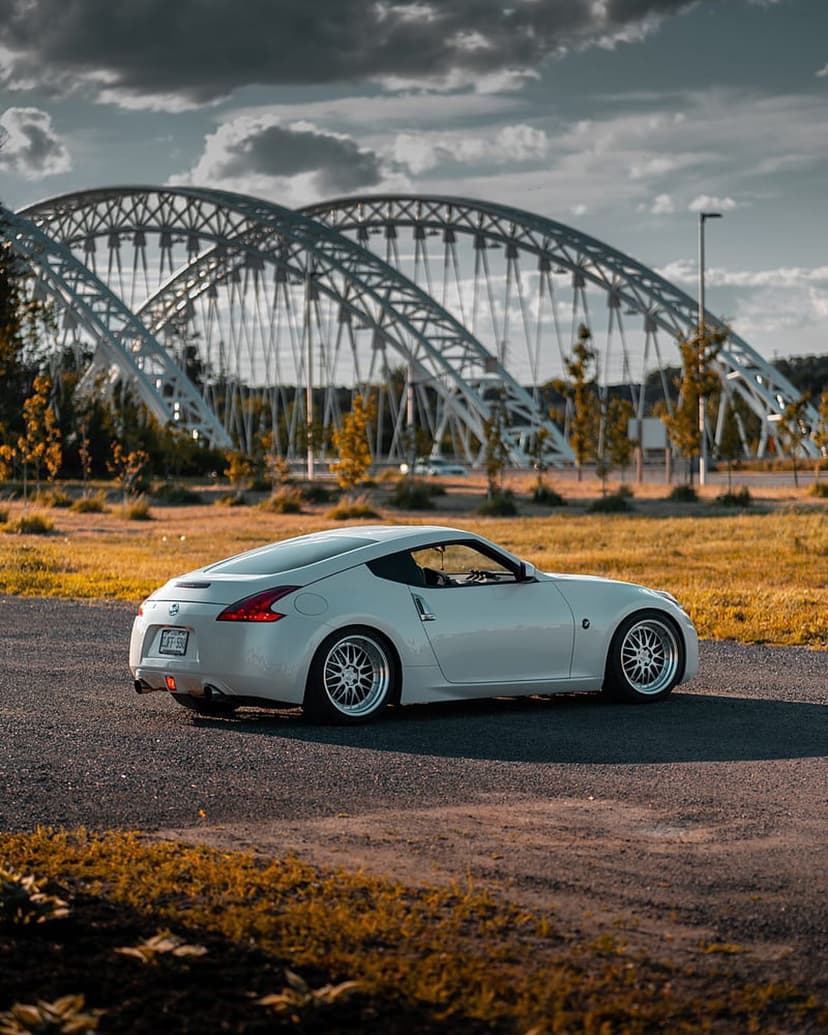 White Nissan 370Z sports car on an asphalt road with a massive arch bridge and golden sunset in the background.