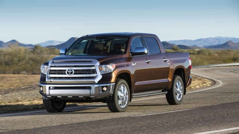 Brown Toyota Tundra pickup truck driving on a winding desert road under a clear blue sky with mountains behind.