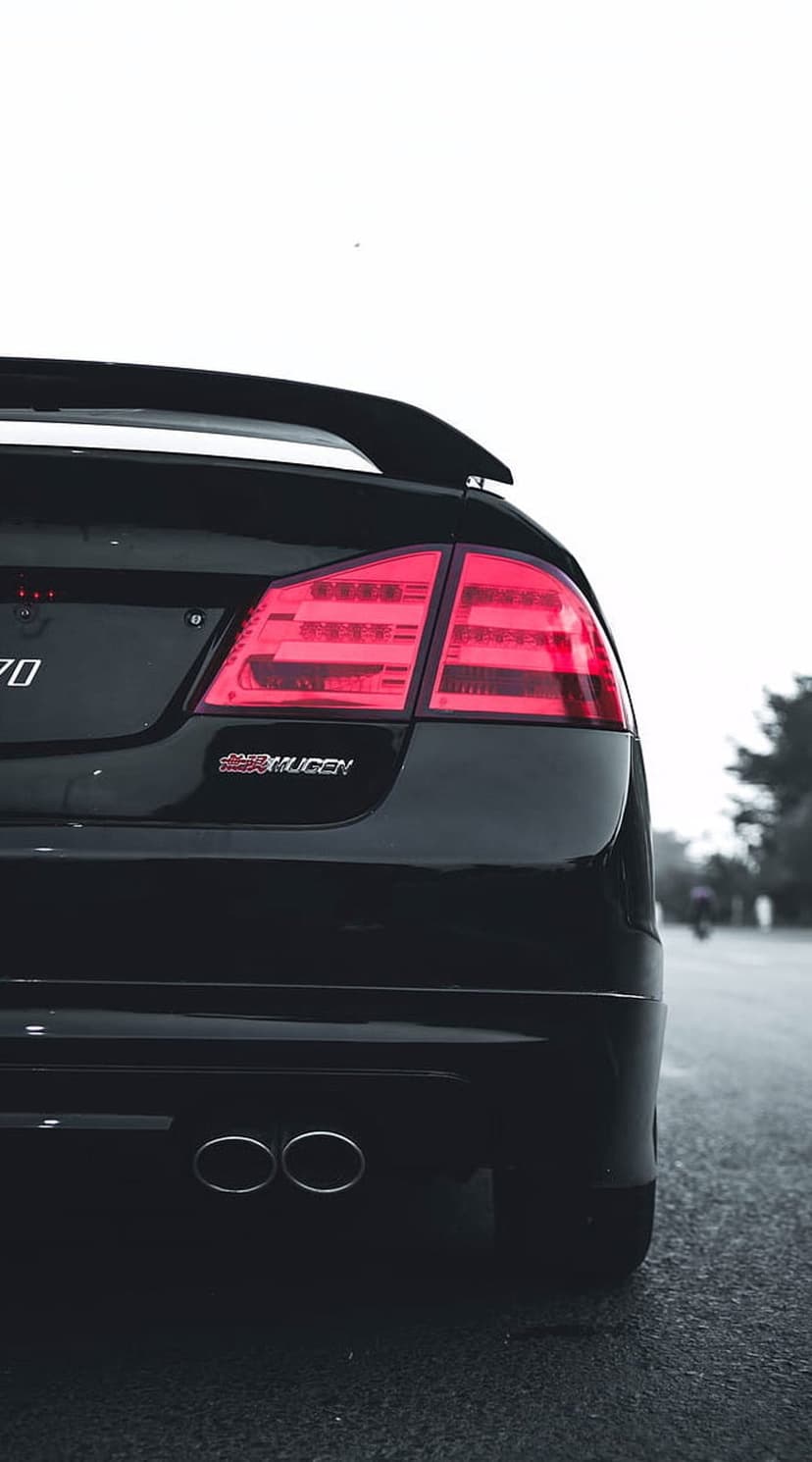 Black Honda Civic rear view with Mugen badge, raised spoiler, dual exhaust pipes, and glowing red taillights.
