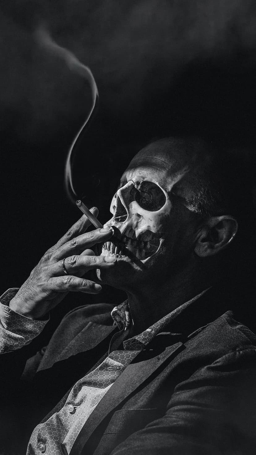 Man with a skull face wearing a suit and tie smokes a cigarette as wisps of gray smoke curl into the dark.