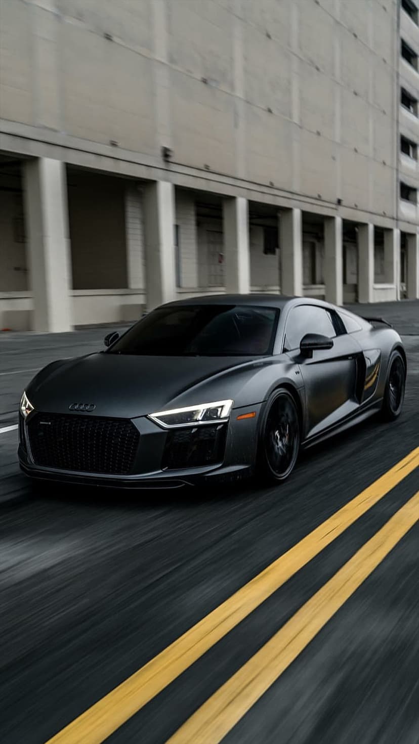 Matte black Audi R8 supercar speeding on an urban road with blurred yellow lines and modern city buildings.