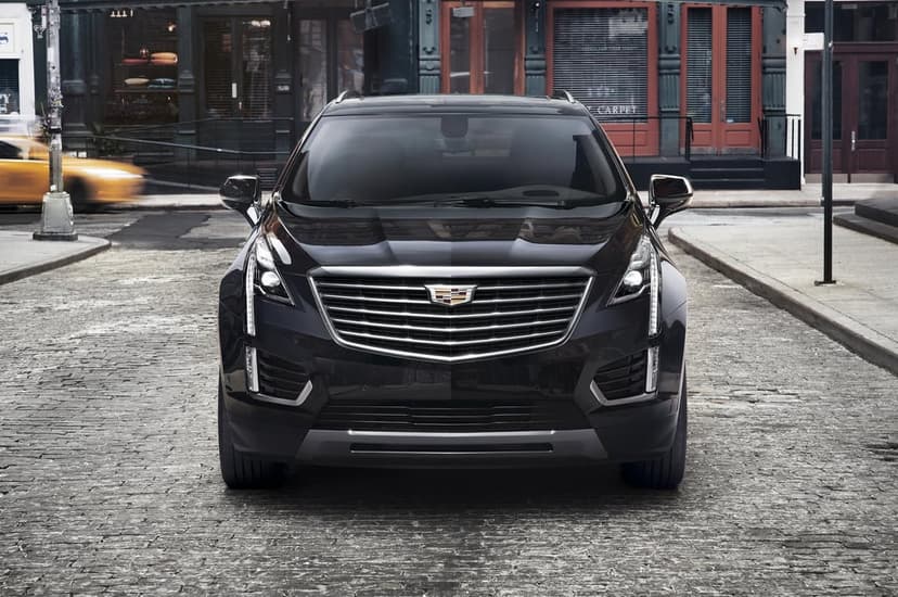 Black Cadillac SUV parked on a grey cobblestone city street with blurred buildings and a yellow taxi behind it.