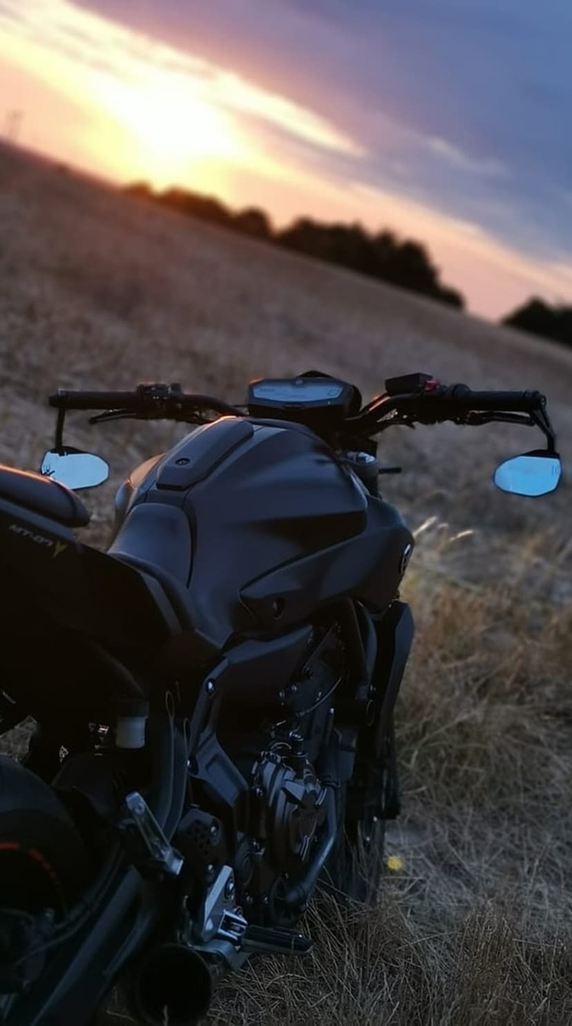 Black motorcycle stands in a dry grassy field beneath a vibrant orange and purple sunset sky during dusk.