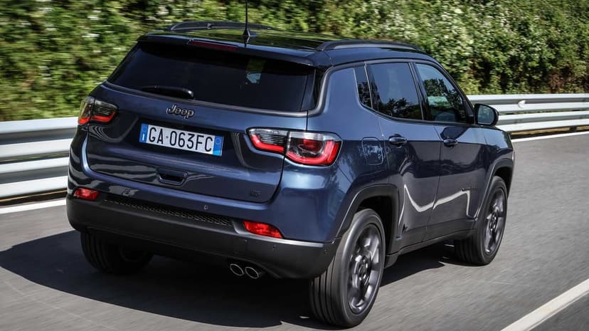 Rear view of a dark blue Jeep Compass driving on an asphalt road with blurred green trees in the background.
