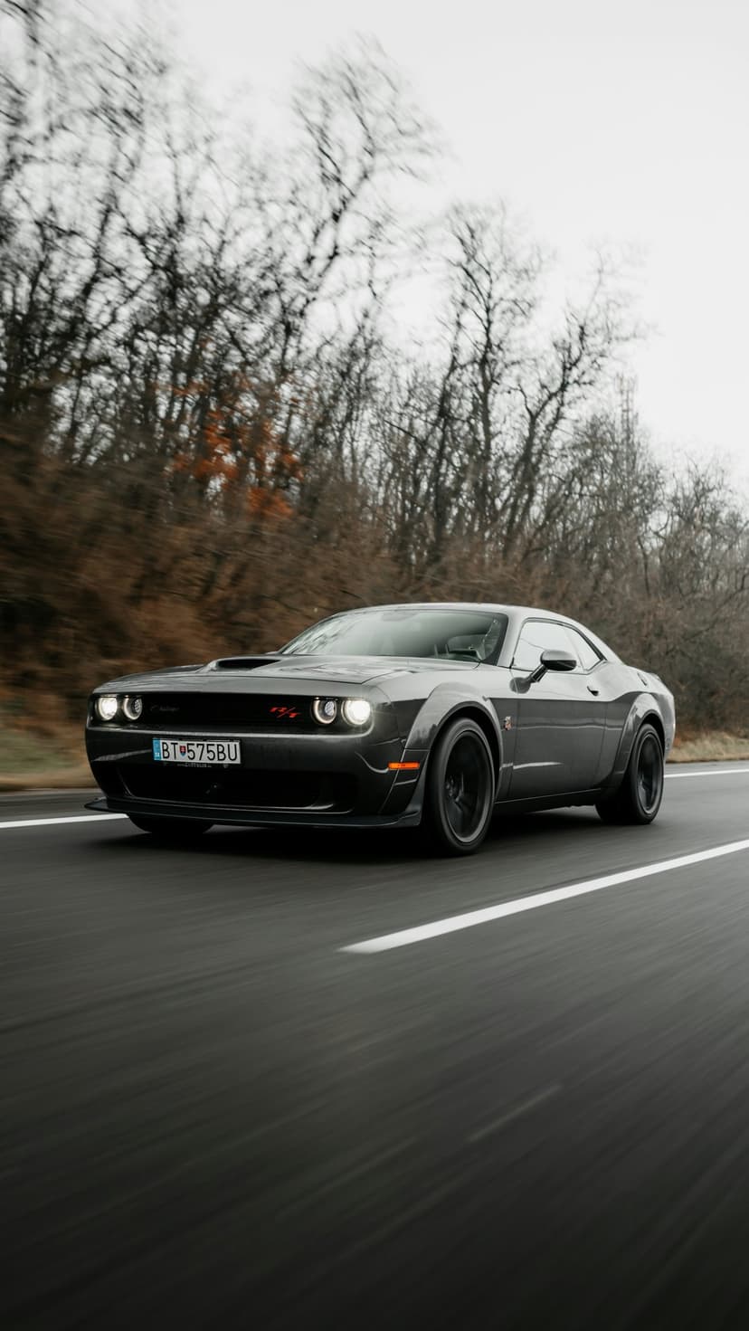 Dark grey Dodge Challenger SRT Hellcat speeding on a paved road lined with blurred autumn trees and foliage.