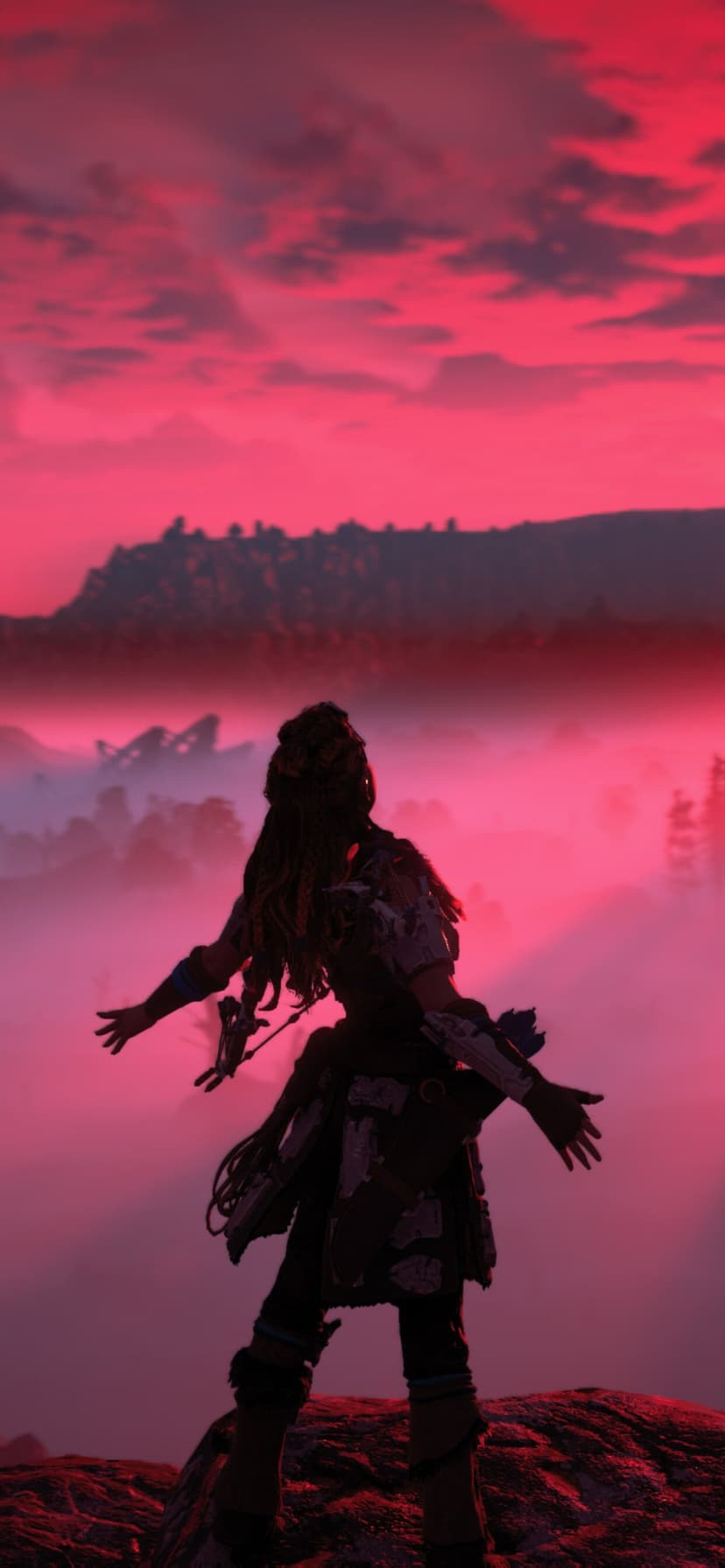 Aloy stands on a rocky ledge with arms outstretched overlooking a misty pink and crimson mountain landscape at dawn.
