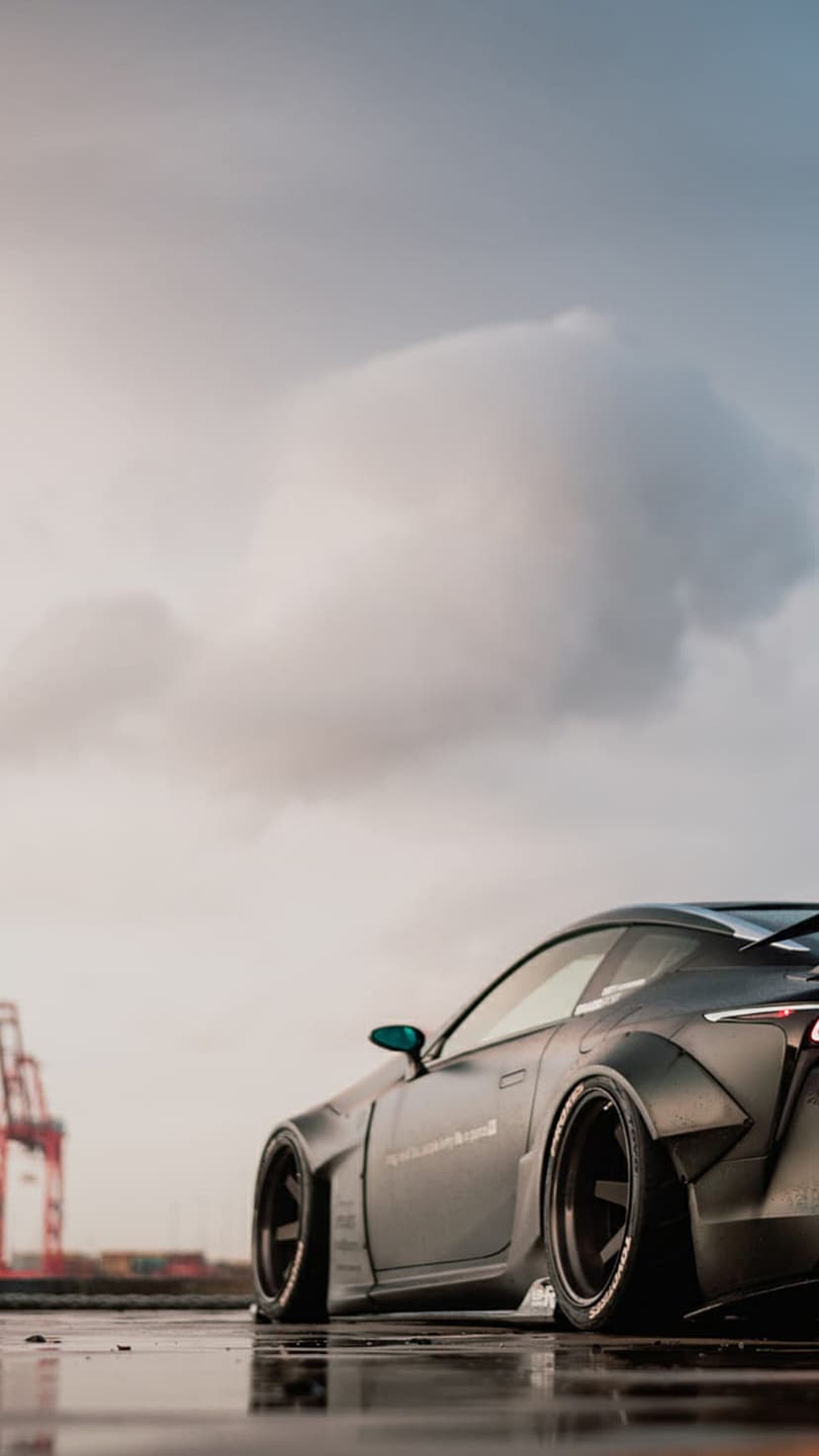 Matte black Lexus LC 500 widebody on wet pavement under a moody sky, showing industrial background reflections.