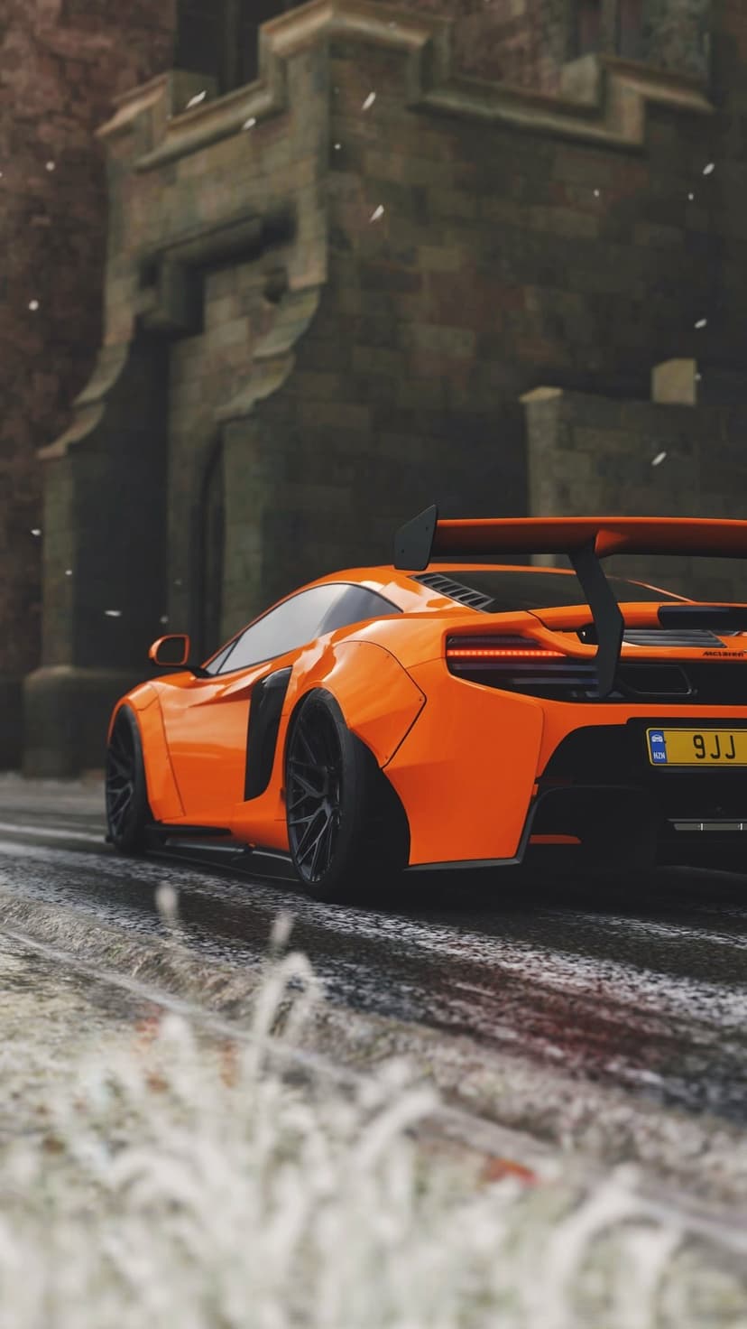 Orange McLaren supercar with a rear spoiler driving on a wet snowy road past ancient stone buildings.