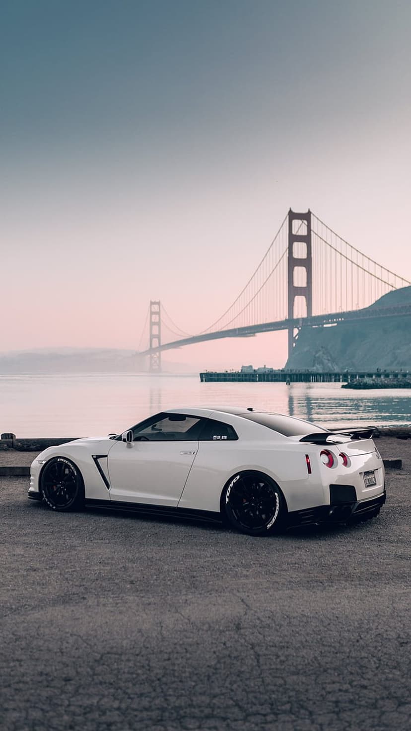 White Nissan GT-R sports car parked on a roadside with the red Golden Gate Bridge visible through morning mist.
