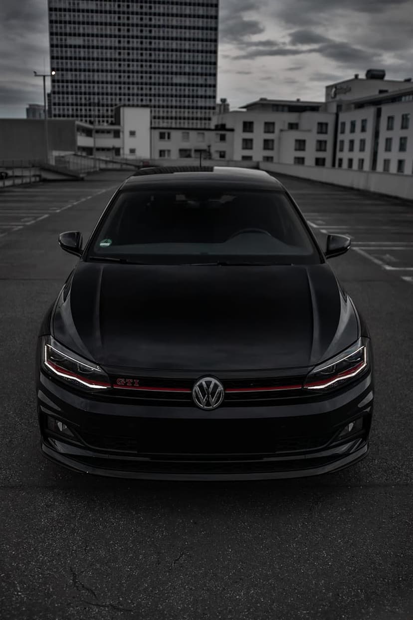Black VW Polo GTI with glowing LED headlights parked in a dark urban lot under a cloudy sky from a low angle.