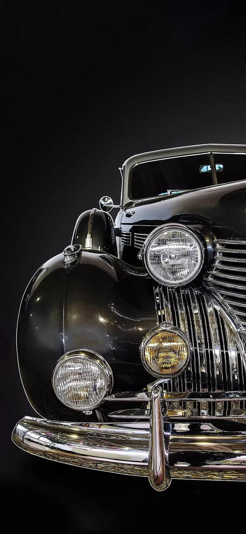 Round glass headlights and a reflective chrome grille on a black vintage car with high-contrast studio lighting.