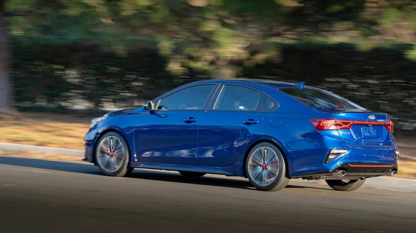 Blue Kia Forte sedan driving fast with motion blur on wheels and a blurred green forest background.