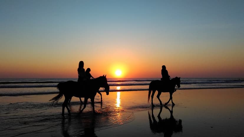 Two horseback riders move along a wet beach as the orange sunset reflects in the calm ocean water and sand.