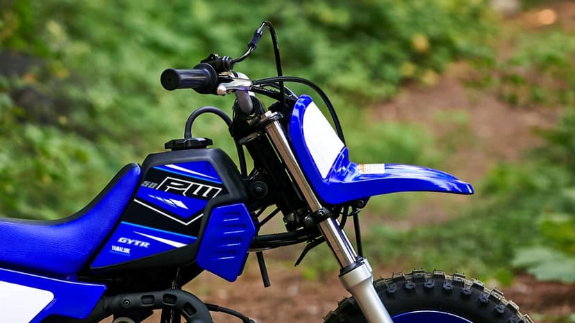 Blue Yamaha PW50 mini dirt bike front view with knobby tires and handlebars against a blurred forest background.
