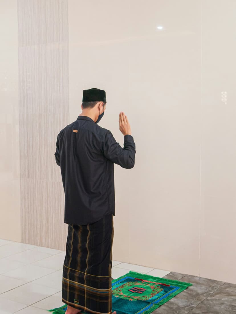 Man in a black cap and plaid sarong stands on a green prayer mat against a tiled wall in a bright mosque interior.