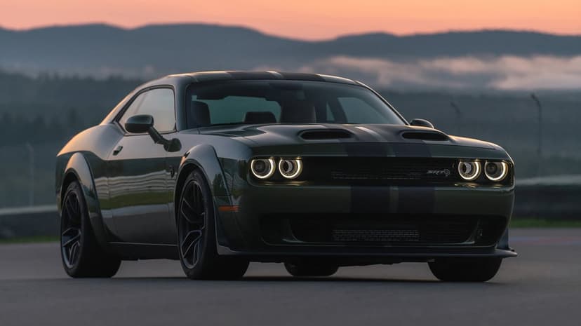 Dark green Dodge Challenger SRT Widebody with glowing halo headlights on a track at sunset with misty mountains.