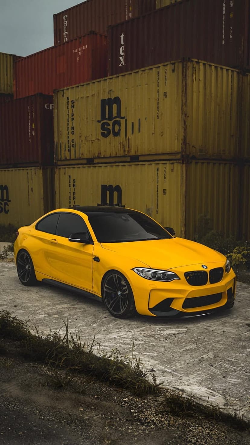 Yellow BMW M2 Coupe with a black roof parked on concrete in front of stacked red and yellow shipping containers.