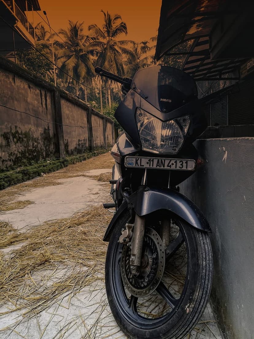 Hero motorcycle in black sits by a rustic wall under green palms and a glowing sunset sky background.