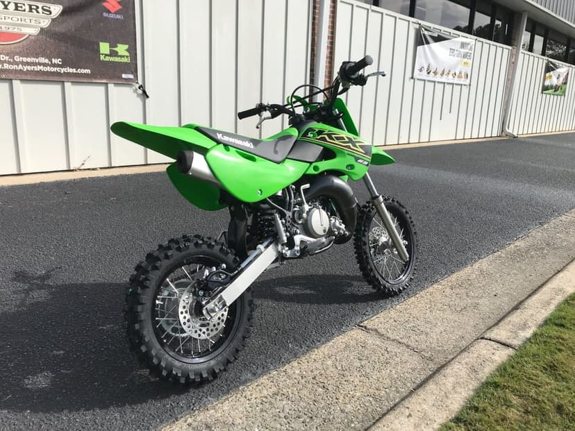 Kawasaki KX 65 dirt bike with lime green bodywork parked on asphalt near a corrugated metal building.