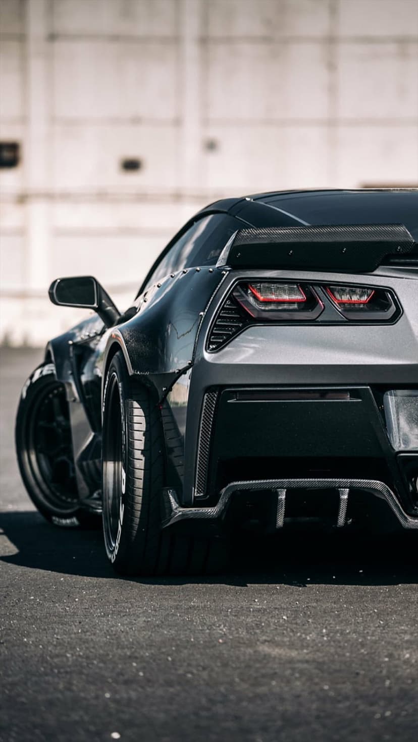 Widebody Corvette Stingray rear with flared fenders and custom spoiler against a blurred industrial backdrop.