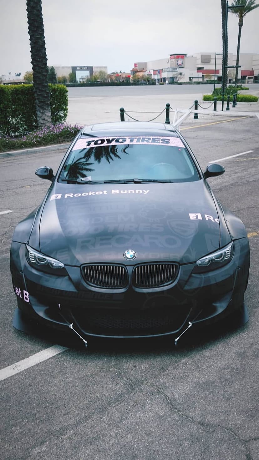Matte black BMW M3 shows aggressive fender flares, a Toyo Tires banner, and racing logos on the hood in front view.