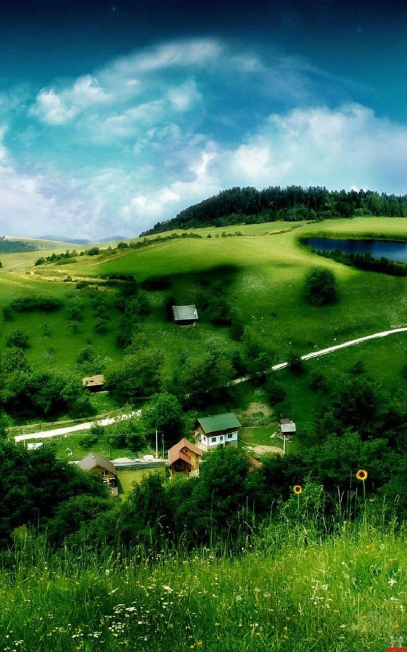 Rolling green hills with small white houses lead toward a calm lake under a bright blue sky with white clouds.