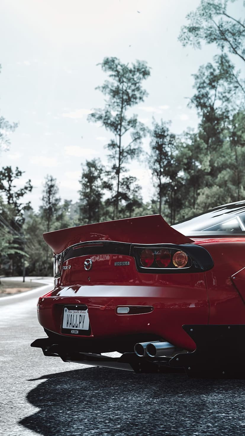 Red Mazda RX-7 with a large GT wing parked on a tree-lined road showing the rear bumper and dual exhaust pipes.