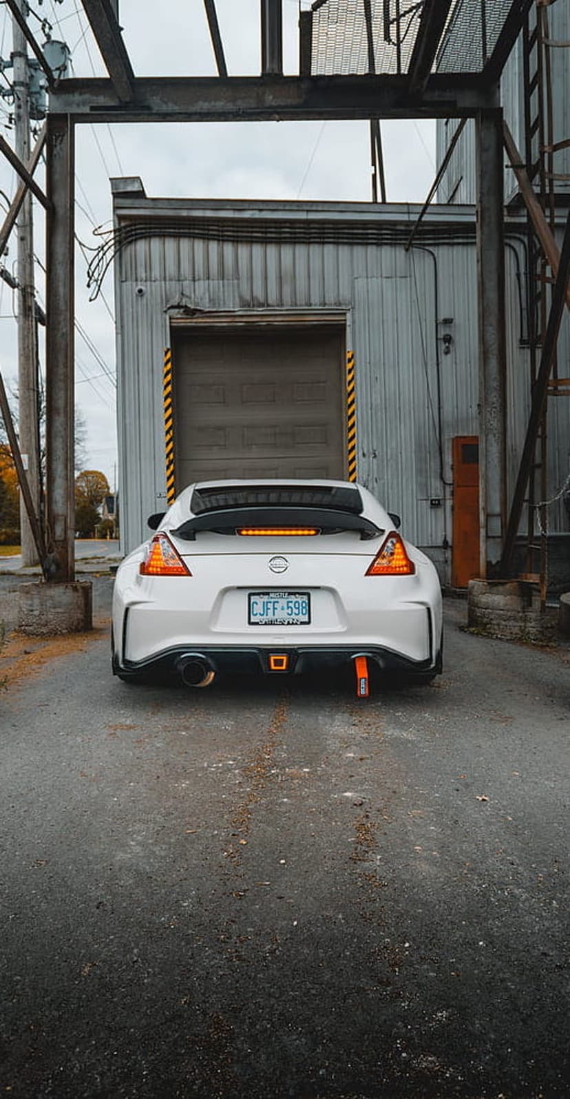 White Nissan 370Z with a rear spoiler and orange tow hook parked on wet asphalt in a dark industrial alleyway.