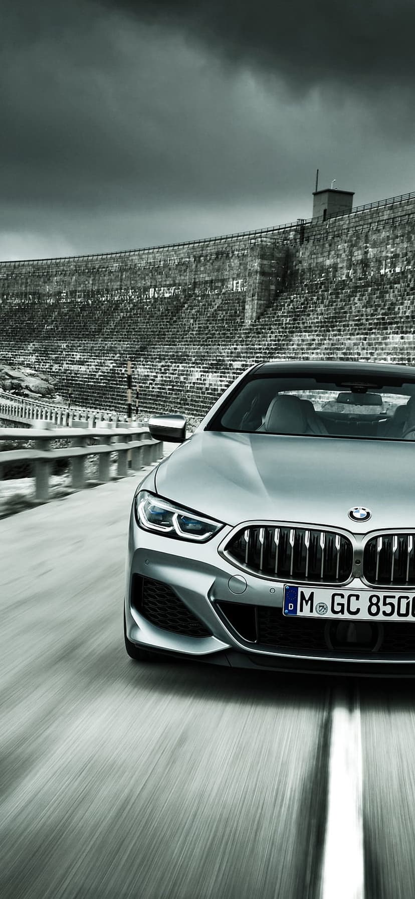 Silver BMW 8 Series convertible speeds along a road with a massive curved concrete dam wall in the background.