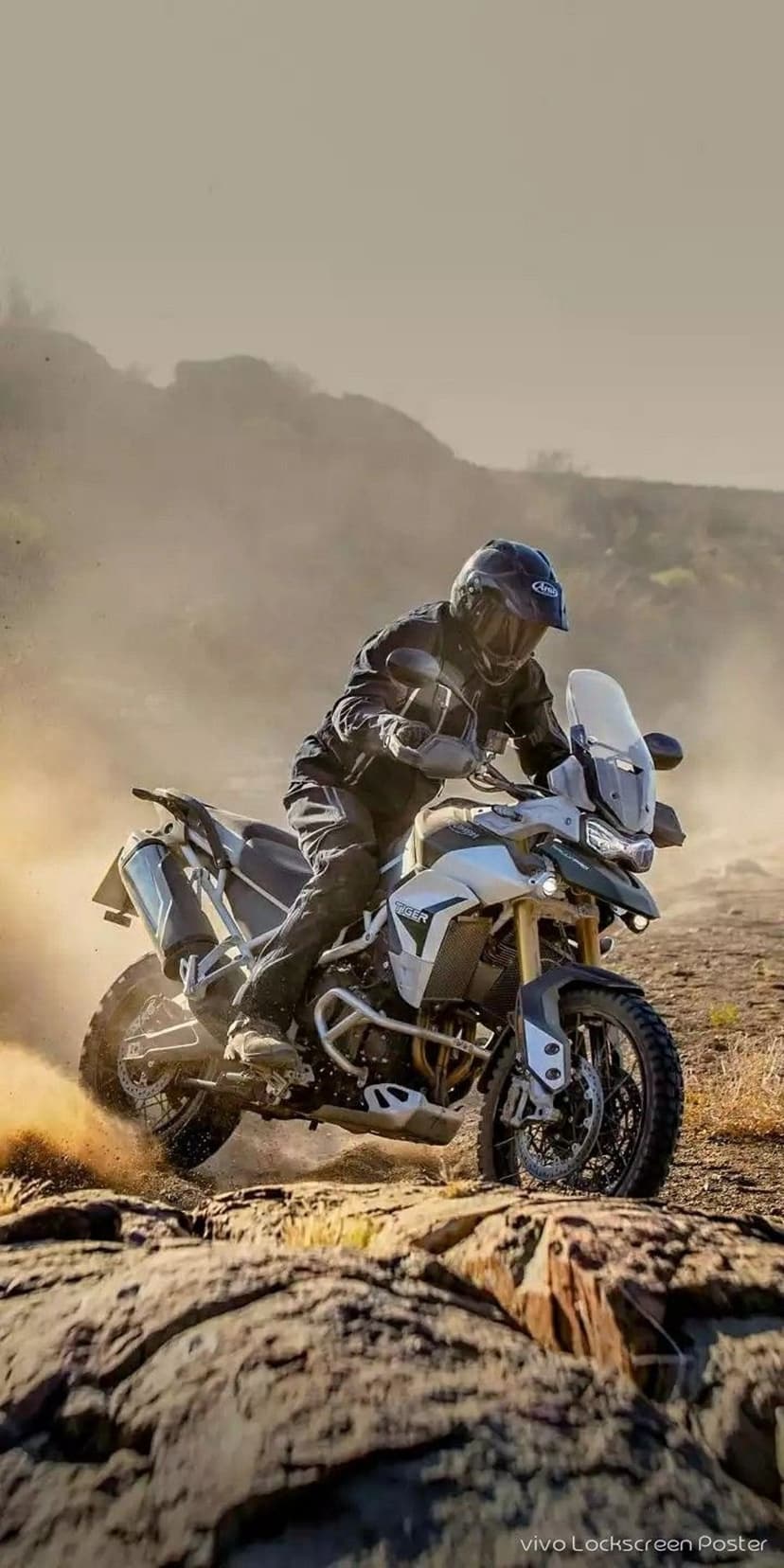 Motorcyclist on a white and green Triumph Tiger bike kicks up dust while riding across a rocky desert landscape.