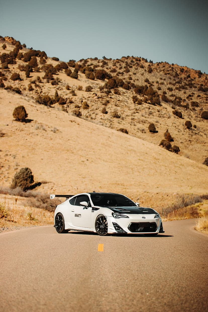White sports car with a black hood driving on a curved desert road under a clear blue sky during the day.