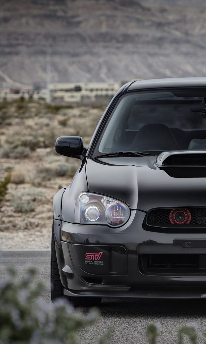 Black Subaru WRX STI front view with hood scoop in a dry desert valley under warm sunset lighting conditions.