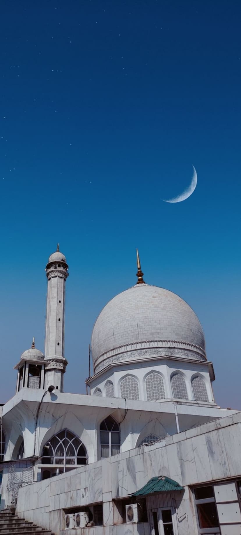 White mosque dome and minaret stand under a glowing crescent moon and starry blue sky in a vertical composition.