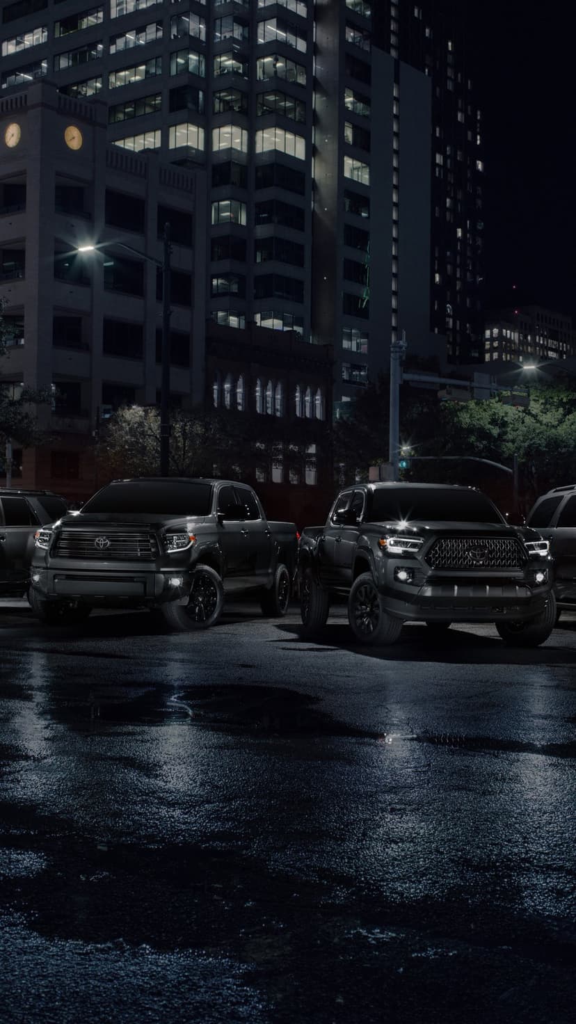 Two black Toyota pickup trucks parked on a wet city street at night under glowing office building lights.