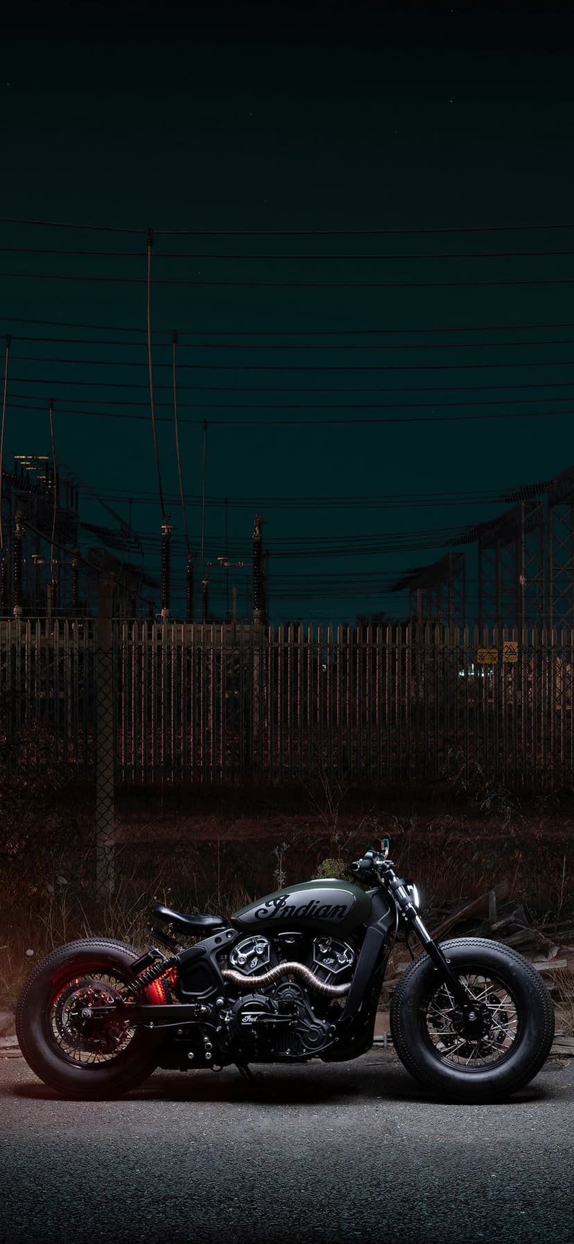 Black Indian motorcycle parked in a dark industrial zone at night with a glowing red taillight and power lines.