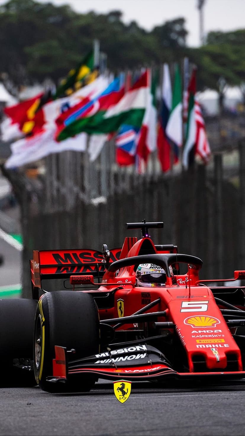 Red Scuderia Ferrari F1 car speeds along a track under fluttering international flags with visible sponsor logos.