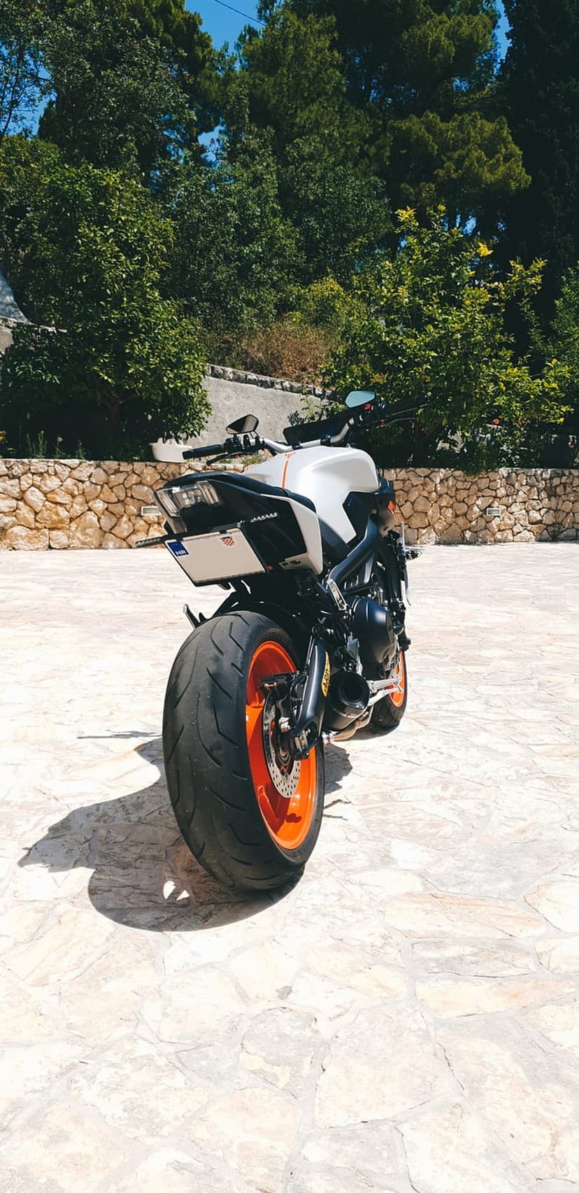 KTM motorcycle rear view showing orange wheels and wide tire on a stone patio under a clear blue summer sky.