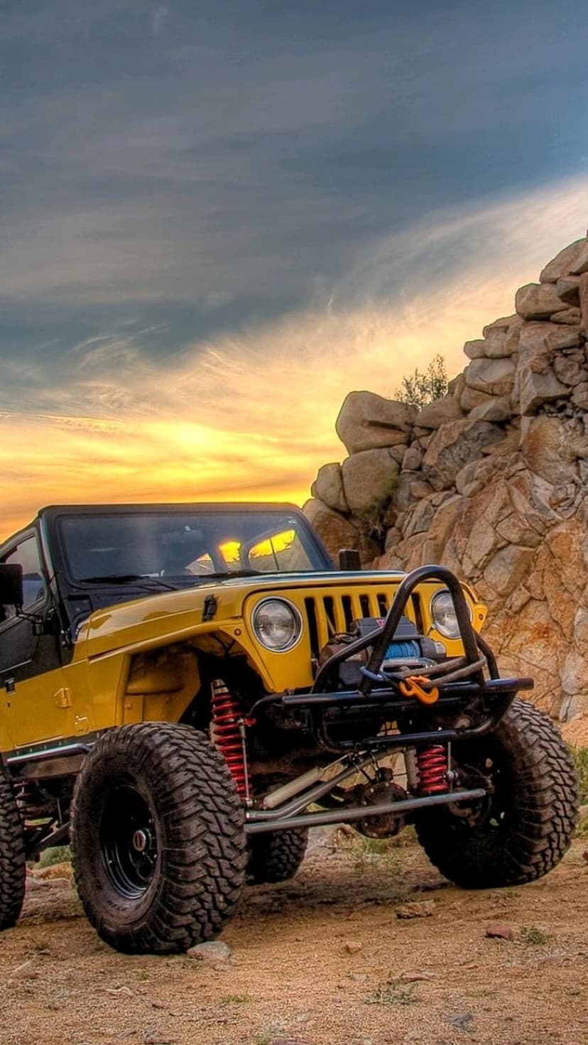 Yellow Jeep Wrangler with oversized tires sits on sharp desert rocks under a glowing orange sunset sky.