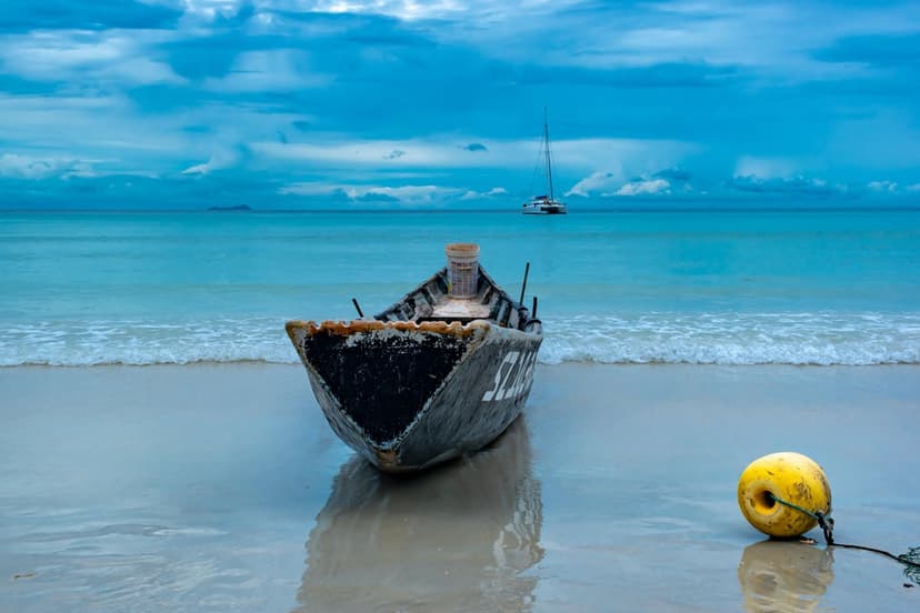 Weathered wooden boat sits on a sandy beach beside turquoise ocean waves under a cloudy sky with a yellow buoy.