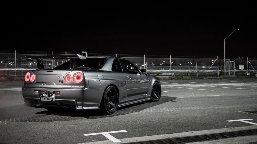 Silver Nissan Skyline GT-R R34 with glowing red taillights parked in a dark lot with industrial fencing.