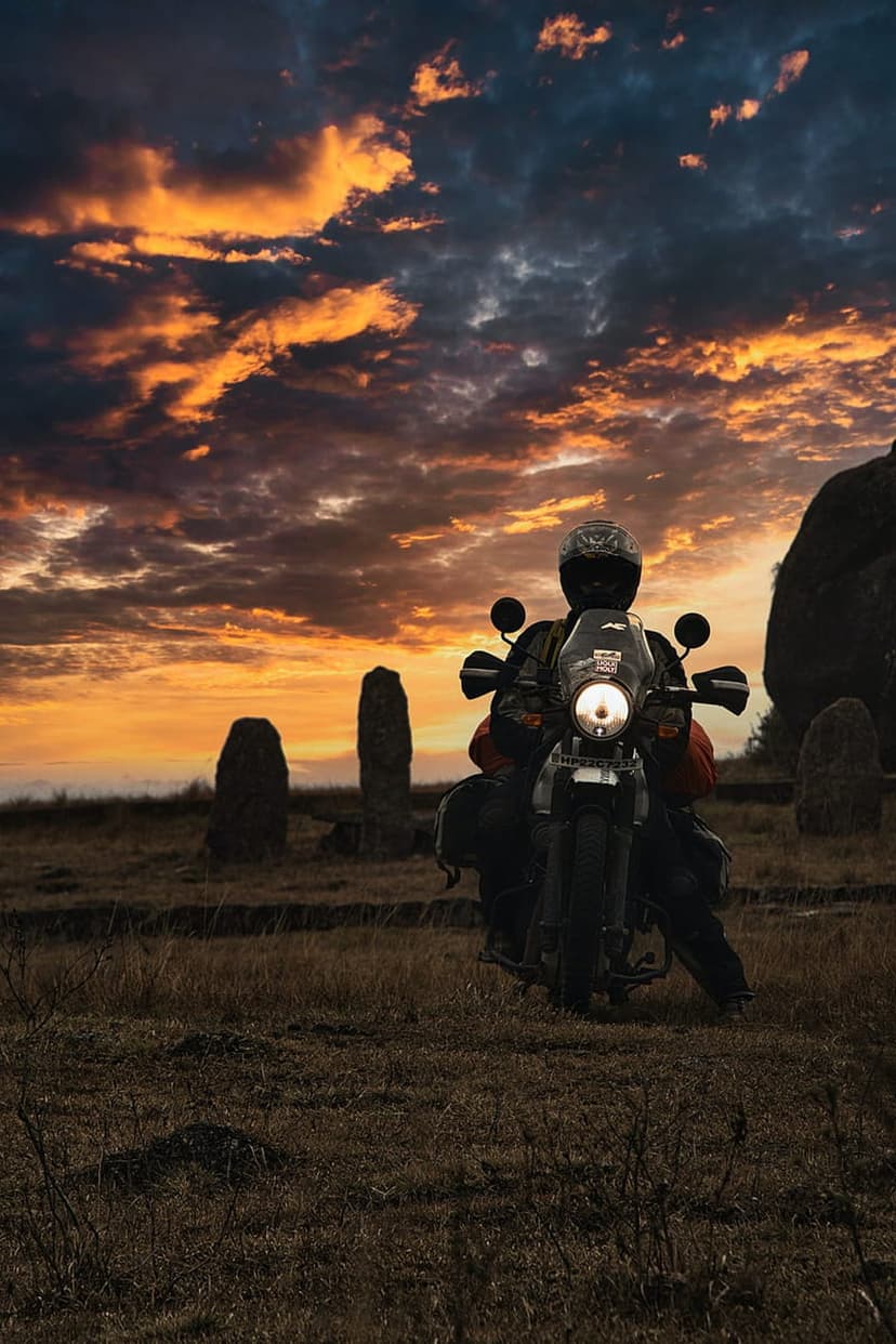 Adventure motorcyclist with luggage stands by tall stone monoliths beneath a burning orange and grey sunset sky.
