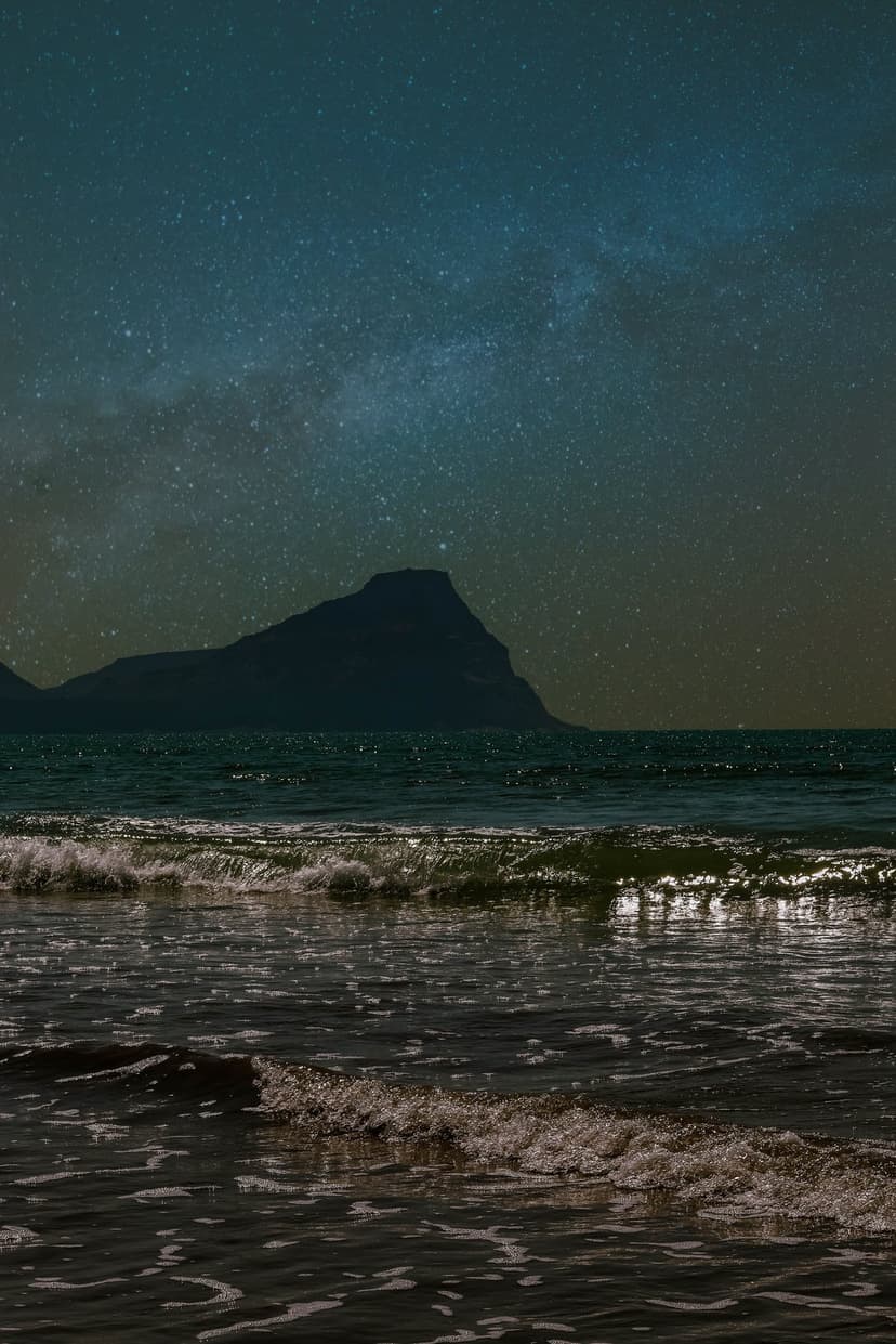 Milky Way galaxy stretching over dark coastal mountains and white waves crashing on a sandy beach at night.