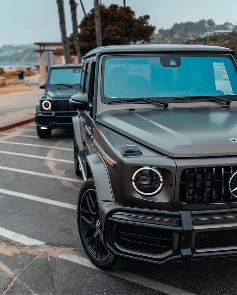 Two dark grey Mercedes G-Wagons parked on a coastal lot near palm trees and the ocean under a grey sky.